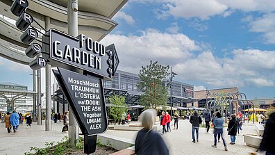 Auf dem Bild ist ein Totempfahl mit der Aufschrift "Food Garden" zu sehen, dahinter stehen Gebäude.