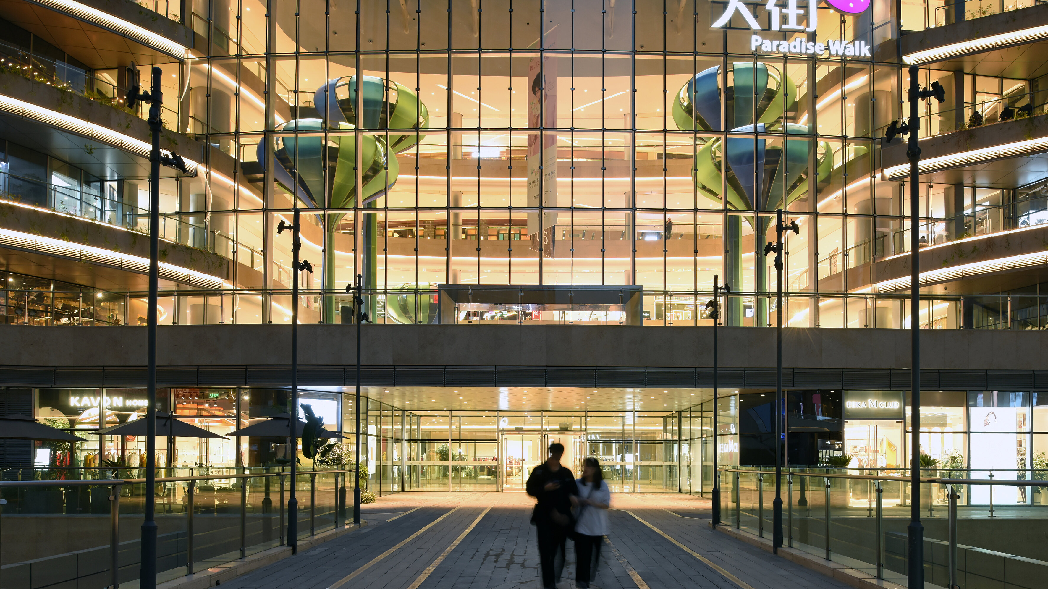 View of the brightly lit entrance area of a shopping centre with a Chinese-language logo on the façade, under which the words 'Paradise Walk' can be read.