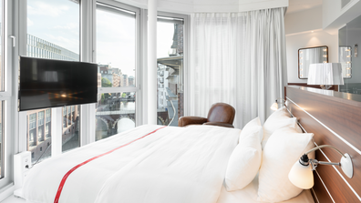 Blick auf ein Schlafbereich in einem Hotelzimmer mit großen lichtdurchfluteten Fenstern.