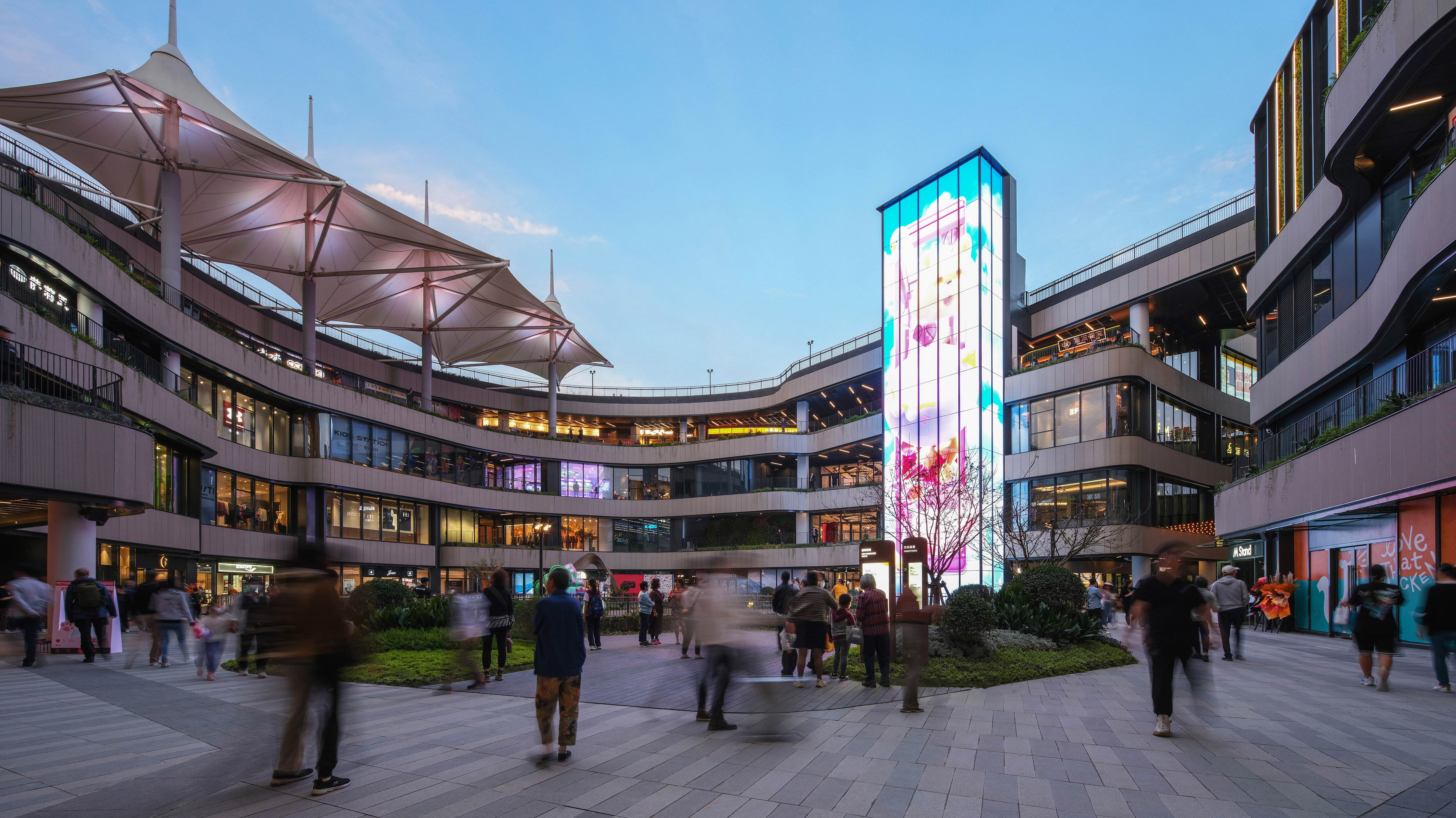 Scene from the outdoor area of a busy shopping centre, characterised by hustle and bustle and a strikingly illuminated pillar.
