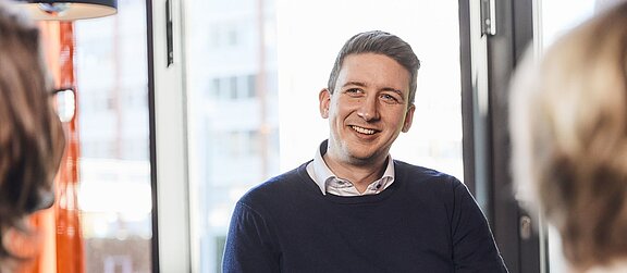 Es ist ein lächelnder Mann von vorne zu sehen. Es werden außerdem zwei Frauenköpfe von hinten angedeutet.