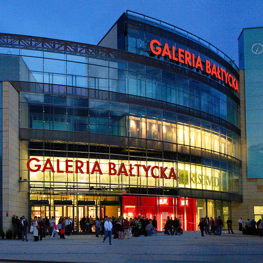 Außenansicht der hell beleuchteten Galeria Bałtyincka, einem modernen, Danziger Einkaufszentrum in der Abenddämmerung. Vor dem Eingang sind Menschen und einige Geschäfte sichtbar.