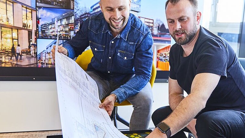 Zwei Personen schauen sich einen Architekturplan auf einem langen weißen Papier an