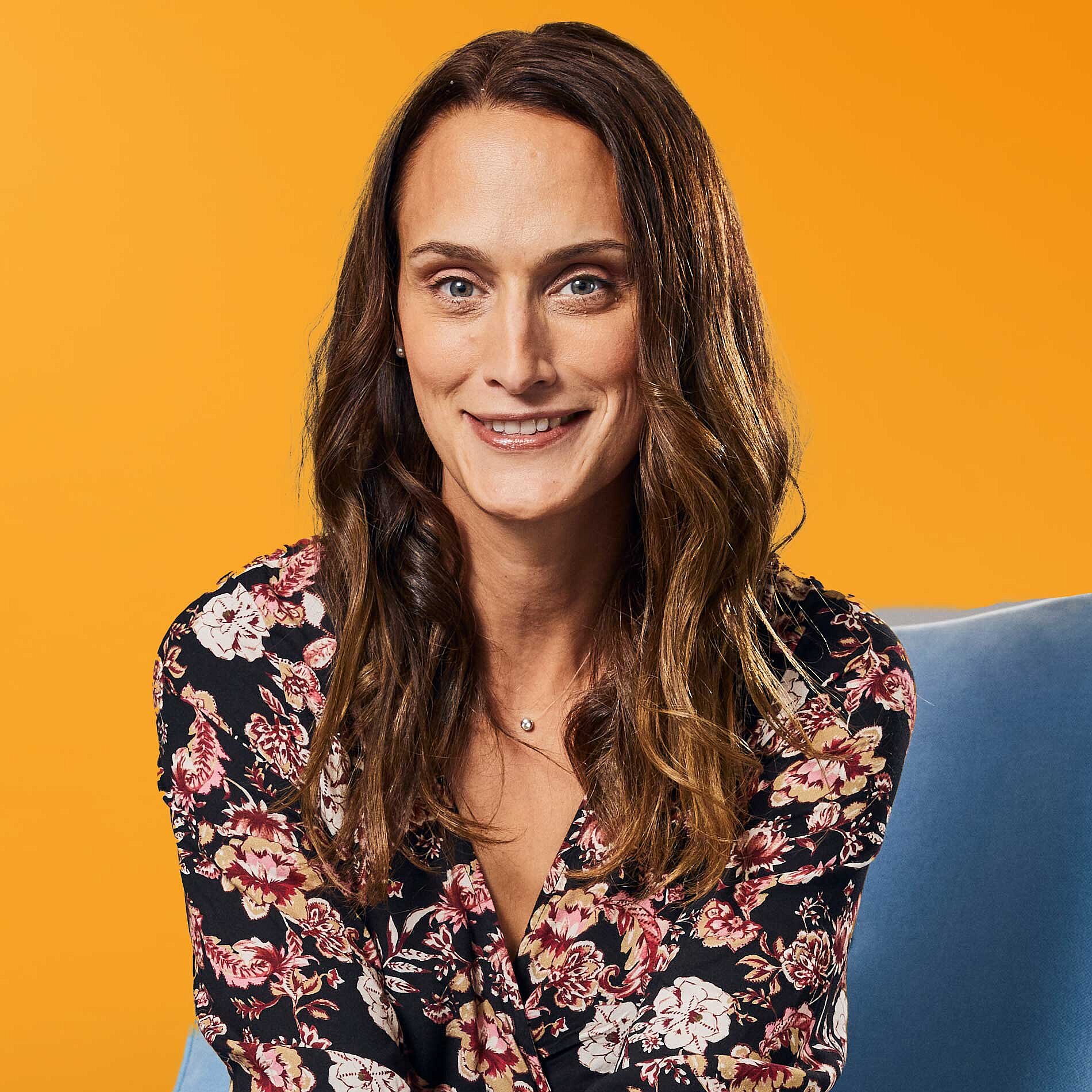 Auf dem Bild ist unsere Kollegin Juliana Schmidt zu sehen. Sie sitzt auf einem blauen Stuhl, vor einem orangenen Hintergrund.
