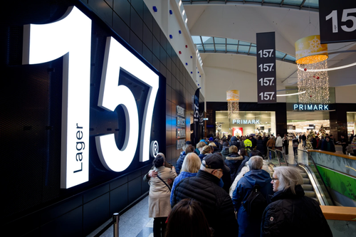 Mehrere Kundinnen und Kunden befinden sich in einem Shopping-Center und stehen vor dem Kleidungsgeschäft Lager 157.