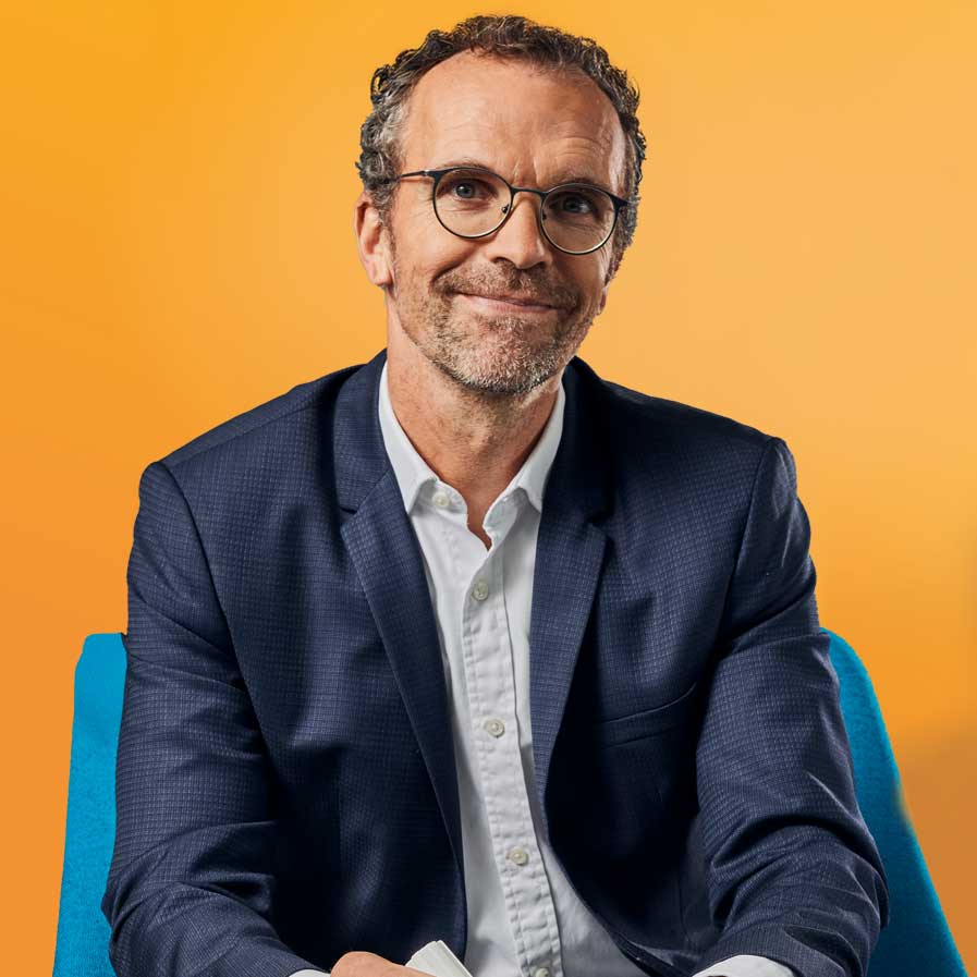 Auf dem Bild ist unser Kollege Martin Alke zu sehen. Er sitzt vor einem orange-farbigem Hintergrund.